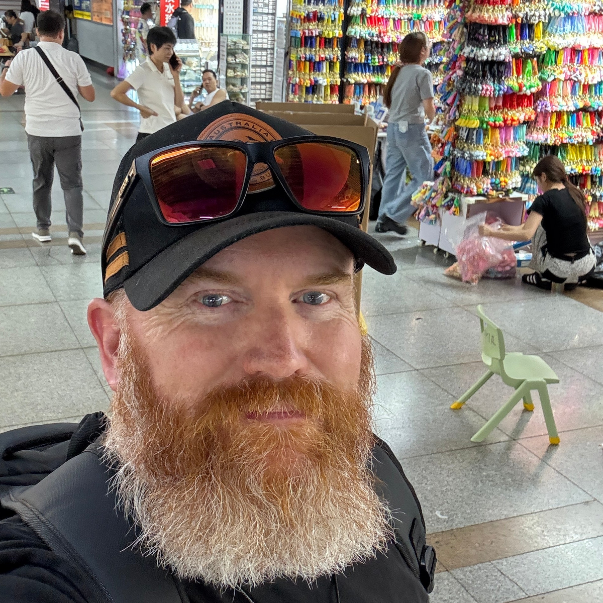 A man with a large red beard takes a selfie in a busy market. He is wearing a black baseball cap with orange-tinted sunglasses resting on top, a black backpack, and a black shirt.